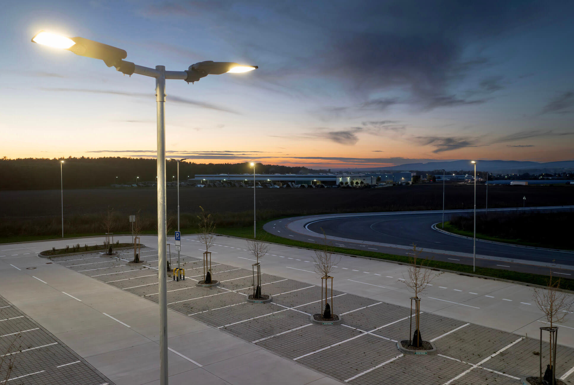 led-parkplatzbeleuchtung-mastleuchte-gewerbe-ledoptix Moderne zweiflammige LED-Mastleuchte erhellt gleichmäßig einen großen Gewerbeparkplatz in der Dämmerung