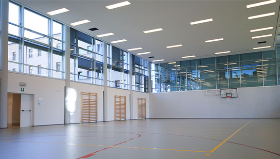 led-sporthallenbeleuchtung-mehrzweckhalle-ledoptix.jpg Blendfreie, gleichmäßige LED-Deckenbeleuchtung in einer modernen Sporthalle mit Basketballkorb und Sprossenwänden